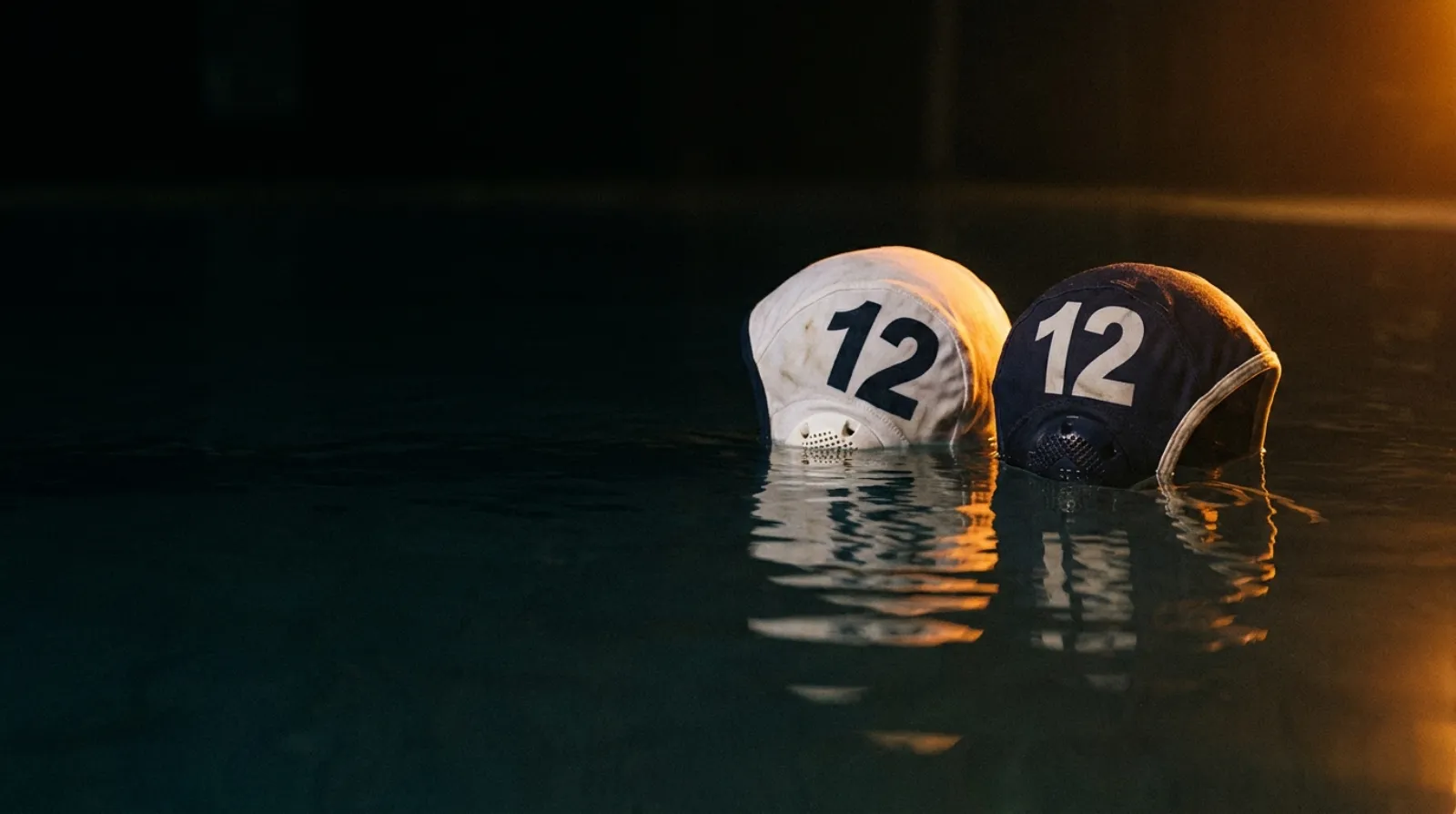 Two water polo caps floating on dark water at night, one white and one dark, both numbered 12.